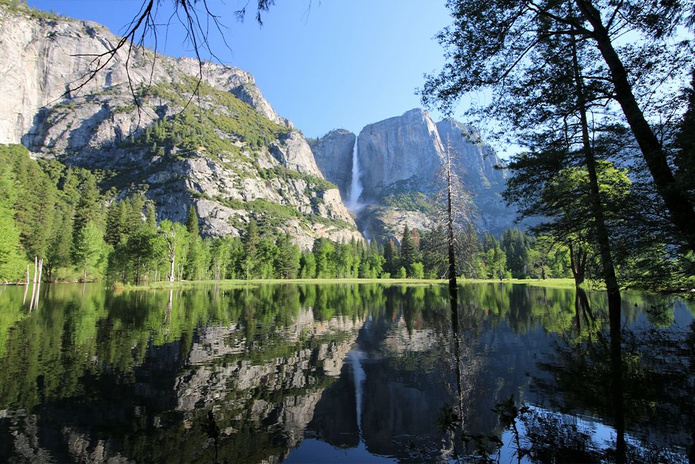 Lake in Yosemite national park with a mighty flowing waterfall cascading down granite rocks. Green trees lining the shoreline of the lake while reflection of the waterfall is seen in the water.